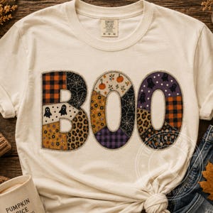 May include: A cream-colored short-sleeve t-shirt featuring the word BOO printed in large letters. Each letter is composed of various patchwork patterns including orange plaid, spiderwebs, ghosts, leopard print, and floral motifs in autumn colors.