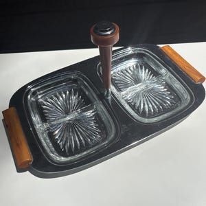 May include: A rectangular chrome serving tray featuring two removable clear glass dishes with a sunburst pattern. The tray includes a central brown handle and two matching brown side handles, providing a structured design for serving food items.