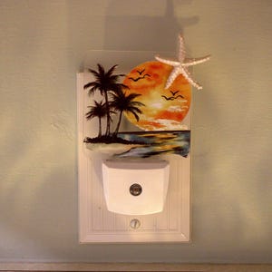 May include: A white plug-in night light featuring a rectangular glass panel with a painted tropical sunset scene. The design includes black silhouettes of palm trees, a bright orange sun, flying birds, and a white starfish attached to the top corner.