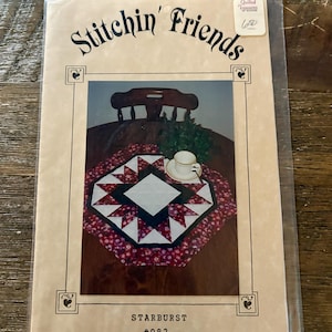 Peut inclure: Un sachet en plastique transparent contenant le patron de courtepointe Stitchin' Friends Starburst #982. La couverture présente une photographie d'un chemin de table en forme d'étoile en tissu rouge, blanc et noir sur une table en bois.