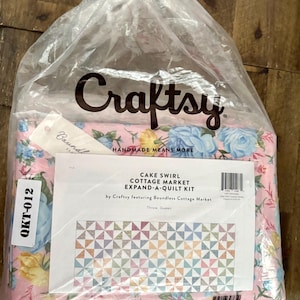 May include: A clear plastic bag containing a Craftsy Cake Swirl Cottage Market Expand-A-Quilt kit. The fabric inside is pink with a floral pattern of blue and yellow roses. A white label displays the text: Cake Swirl Cottage Market Expand-A-Quilt Kit.
