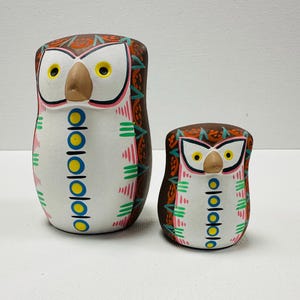 May include: Two owl figurines with brown, white, and orange painted details. The owls feature yellow eyes, a tan beak, and a vertical pattern of blue and yellow circles on their white chests, accented by pink and green geometric markings.