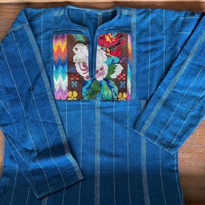 Op de afbeelding: Een blauw shirt met lange mouwen, een verticaal krijtstreepmotief en een centraal rechthoekig paneel op de borst. Dit paneel toont een kleurrijk geborduurd ontwerp van witte bloemen, groene bladeren en een rode roos op een bruine achtergrond.