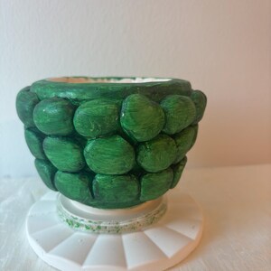 May include: A small, rounded bowl with a textured exterior featuring raised, pebble-like green shapes. The bowl sits on a circular, white, fluted base. The interior of the bowl is white, and the green paint shows visible brushstroke textures.
