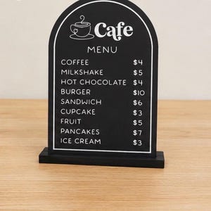 May include: An arched black menu sign with white text and a coffee cup icon. The text reads: CAFE MENU, COFFEE $4, MILKSHAKE $5, HOT CHOCOLATE $4, BURGER $10, SANDWICH $6, CUPCAKE $3, FRUIT $5, PANCAKES $7, ICE CREAM $3. It stands on a black base.