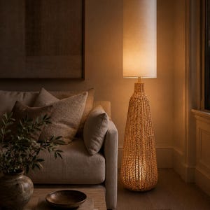 May include: A floor lamp with a tall, cylindrical off-white fabric shade and a base crafted from woven natural fibers in a tapered, bottle-like shape. The lamp emits a warm, soft glow, illuminating the surrounding area with a gentle light.