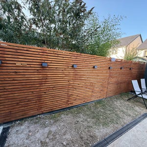 May include: A tall, horizontal slat fence made of stained brown wood, featuring a series of small, rectangular black wall-mounted lights spaced evenly along the upper section. The fence is constructed from thin, parallel wooden boards.