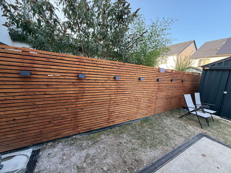 May include: A tall, horizontal slat fence made of stained brown wood, featuring a series of small, rectangular black wall-mounted lights spaced evenly along the upper section. The fence is constructed from thin, parallel wooden boards.