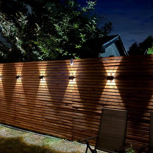 May include: A series of seven rectangular black outdoor wall lights mounted on a horizontal wooden slat fence. Each fixture emits a bright, warm white light in an upward and downward X-shaped pattern, illuminating the dark brown wood texture.