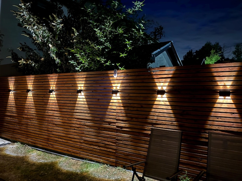 May include: A series of seven rectangular black outdoor wall lights mounted on a horizontal wooden slat fence. Each fixture emits a bright, warm white light in an upward and downward X-shaped pattern, illuminating the dark brown wood texture.