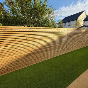 May include: A tall, horizontal slat fence constructed from light brown natural wood. The fence features thin, evenly spaced wooden boards running parallel to each other, creating a clean, linear pattern that spans the length of the outdoor area.