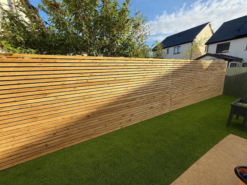 May include: A tall, horizontal slat fence constructed from light brown natural wood. The fence features thin, evenly spaced wooden boards running parallel to each other, creating a clean, linear pattern that spans the length of the outdoor area.