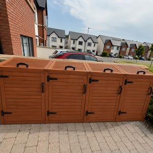 May include: A large, rectangular, four-section wooden storage unit with a slatted design. Each section features a hinged lid with a black handle and a front door with black hinges and a black handle, providing a secure enclosure for outdoor items.