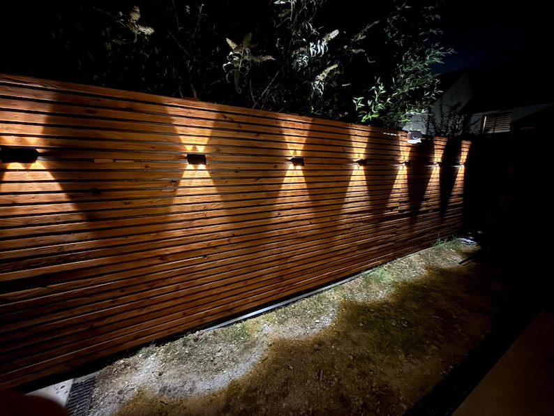 May include: A horizontal wooden slat fence featuring multiple black wall-mounted light fixtures. Each fixture emits a dual-directional beam of warm light, creating an X-shaped pattern against the brown wood grain of the fence panels at night.