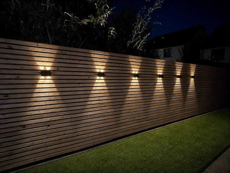 May include: A horizontal wooden slat fence featuring six rectangular wall-mounted light fixtures. Each fixture emits a bright, warm white light in an upward and downward X-shaped pattern against the natural brown wood grain of the fence panels.