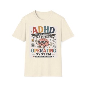 May include: A cream-colored short-sleeve t-shirt featuring a graphic of a cartoon brain wearing sunglasses. The text reads: ADHD IT'S NOT A DISORDER IT'S A DIFFERENT OPERATING SYSTEM SOME UPDATES TAKE FOREVER, accompanied by icons of a lightbulb and paper plane.