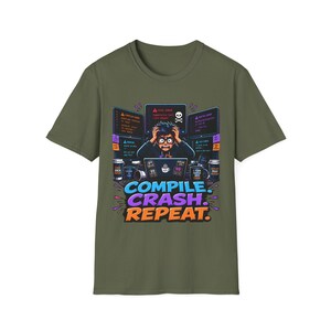 May include: An olive green t-shirt featuring a graphic of a stressed person at a computer with multiple screens displaying error messages and a skull icon. Below the illustration, the text reads: COMPILE. CRASH. REPEAT. in blue, purple, and orange.