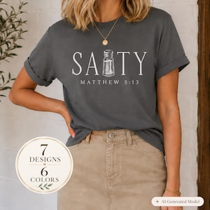 May include: A dark grey short-sleeve t-shirt featuring the white text SALTY with a salt shaker graphic replacing the letter L and the text MATTHEW 5:13 printed underneath. The shirt has a crew neckline and rolled sleeves, paired with a tan skirt.