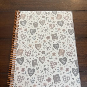 May include: A spiral-bound notebook featuring a repeating pattern of hearts, flowers, bows, and books with crosses. The hearts contain the text "Jesus loves you" in a dark font. The design uses a muted color palette of gray, brown, and white.