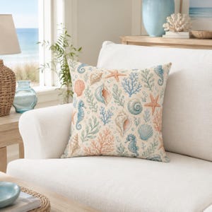 Op de afbeelding: Een vierkant sierkussen met een herhalend patroon van zeepaardjes, zeesterren, diverse schelpen en koraaltakken in blauwe, oranje en beige tinten op een lichtcrème achtergrond. Het ontwerp bevat zeewier en kleine belletjes.