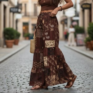 Pode incluir: Uma saia comprida castanha com um design de patchwork que apresenta vários padrões em tons de castanho escuro e creme. A saia tem uma cintura alta elástica com cordão e uma silhueta em camadas que flui em direção à bainha.
