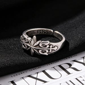 May include: An adjustable silver ring featuring an oxidized finish and an embossed star and cross design. The inner band is clearly stamped with the text S925, indicating the sterling silver composition of this open-style band ring.
