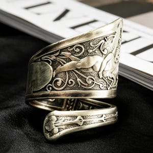 Puede incluir: Un brazalete ancho de tono plateado con un intrincado relieve de una figura alada rodeada de volutas ornamentales. El metal tiene un acabado oscuro y antiguo con un diseño de espiral que se estrecha hacia un extremo redondeado.