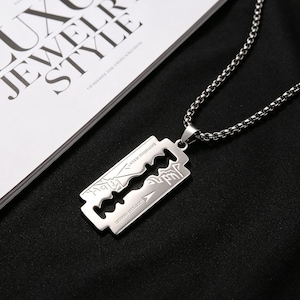 May include: A silver-toned stainless steel pendant shaped like a razor blade, featuring intricate etched line art designs and text, suspended from a matching silver-toned metal box chain necklace with a polished finish and a secure link construction.