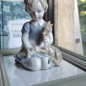 Może przedstawiać: Figurka z glazurowanej porcelany przedstawiająca dziecko o jasnobrązowych włosach, ubrane w niebieską sukienkę z białym kołnierzykiem, klęczące i trzymające małego brązowego jelonka. Figurka ma błyszczące wykończenie w odcieniach błękitu, bieli i brązu.
