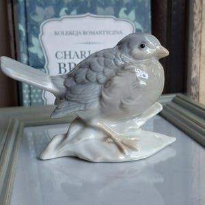 Op de afbeelding: Een porseleinen vogelbeeldje met een glanzende witte en zachtgrijze afwerking. De vogel zit op een kleine, getextureerde basis met lichtbruine accenten op de poten. De vogel heeft een glad, rond lichaam, een puntige snavel en een lange staart.