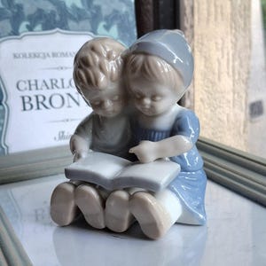 Może przedstawiać: Porcelanowa figurka przedstawiająca dwoje dzieci siedzących razem i czytających otwartą książkę. Dzieci mają jasnobrązowe włosy, a postać po prawej stronie ma na sobie niebieski czepek i niebieską sukienkę. Figurka ma błyszczące wykończenie w odcieniach niebieskiego i beżu.