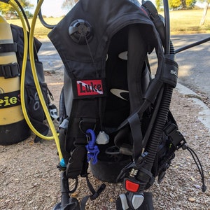 Pode incluir: Colete equilibrador (BCD) de mergulho preto com equipamento acoplado. Apresenta um patch "Luke" vermelho e branco, uma mangueira corrugada e um regulador. Um cilindro de ar amarelo é visível no fundo.