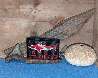Könnte beinhalten: Ein rechteckiges schwarzes Stoffband mit einer gestickten rot-weißen Haifisch-Silhouette über dem in rotem Garn gestickten Namen Anika. Das Design ist mittig auf dem strukturierten schwarzen Gurtband platziert und erzeugt einen klaren Kontrast.