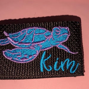 May include: A rectangular black woven fabric patch featuring an embroidered sea turtle in bright blue with a purple outline. The name Kim is embroidered in bright blue cursive script in the lower right corner of the patch surface.