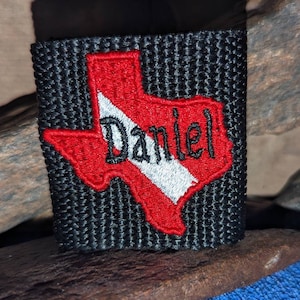 May include: Black embroidered patch featuring the outline of Texas in red with a white stripe and the name "Daniel" in black script. The patch is on a textured black background.