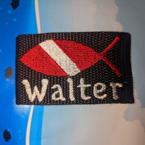 May include: A rectangular black embroidered patch featuring a red and white fish-shaped diver down flag symbol above the white embroidered text Walter. The patch has a textured fabric surface with visible stitching throughout the design.