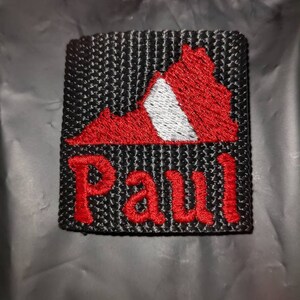 May include: A square black patch featuring an embroidered red and white map shape of Virginia at the top. Below the map, the name Paul is embroidered in red thread. The patch has a textured, woven black background with visible stitching throughout.