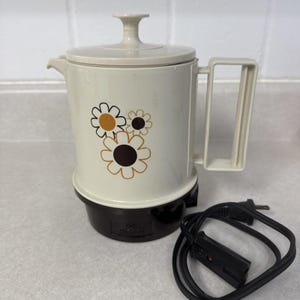 May include: A cream-colored electric percolator with a lid and a side handle. The body features a graphic print of three stylized flowers in brown and mustard yellow. The base is dark brown and includes a detachable black power cord with a plug.