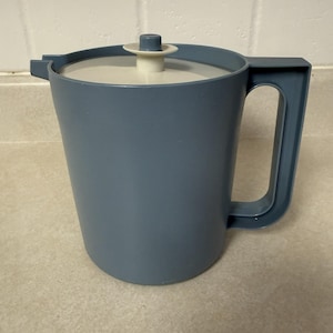 May include: A slate blue plastic pitcher with a matching handle and a spout. The pitcher features a circular, off-white lid with a small, central knob for lifting. The design is simple and functional, with a smooth, matte finish throughout the body.