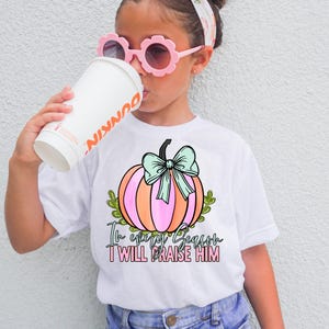Può includere: Una t-shirt bianca a maniche corte con la grafica di una zucca rosa e arancione con un fiocco verde chiaro. Il testo recita In every Season I WILL PRAISE HIM. Il design include foglie verdi che circondano la base della zucca.