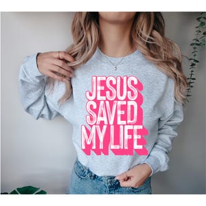 Puede incluir: Una sudadera de cuello redondo gris claro con el texto JESUS SAVED MY LIFE en letras de bloque de color rosa brillante con un efecto blanco desgastado. La prenda de manga larga tiene un corte clásico y se combina con vaqueros azules.