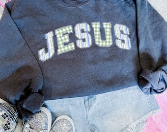 May include: A dark charcoal gray crewneck sweatshirt featuring the word JESUS in large, appliqued letters. The letters alternate between a green and white gingham pattern and a blue and white striped floral pattern, all outlined in white.