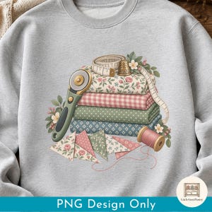 Puede incluir: Una sudadera gris claro de cuello redondo con un gráfico de telas apiladas con estampados florales, de cuadros y de lunares. Un cortador rotativo, una cinta métrica, un dedal, un carrete de hilo rosa y triángulos de tela rodean la pila.