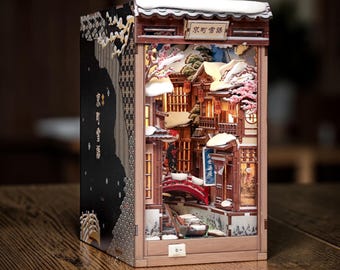 May include: A rectangular wooden book nook diorama featuring a miniature Japanese street scene with snow-covered roofs, a red arched bridge, and illuminated windows. The exterior side panel is black with gold patterns and Japanese text.
