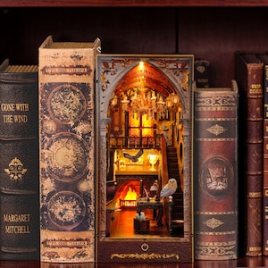 May include: A book nook insert featuring a miniature interior scene with a lit fireplace, a chandelier, a staircase, and an owl figure. The structure is designed to fit between books and includes a power button at the base, creating a warm, illuminated display.