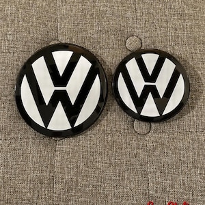 Könnte beinhalten: Zwei kreisförmige Volkswagen-Embleme mit glänzend schwarzem Hintergrund und einem weißen V- und W-Logo. Die Embleme sind rund und flach, wobei eines deutlich größer ist als das andere. Der Text SamStyling ist unten rechts in Rot zu sehen.