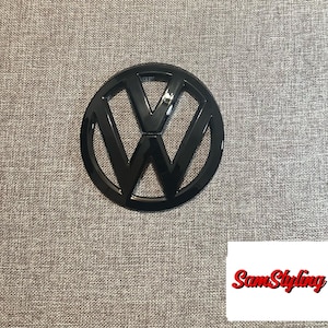 May include: A glossy black Volkswagen emblem featuring the iconic V and W letters enclosed within a circular border. In the bottom right corner, there is a white rectangular label with the red text SamStyling written in a cursive script font.