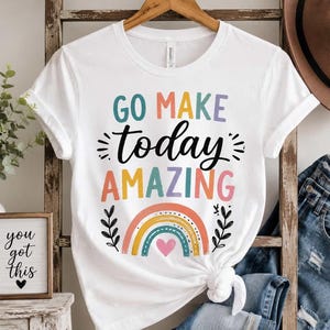 Könnte beinhalten: Ein weißes Kurzarm-T-Shirt mit dem Text GO MAKE today AMAZING in bunten Buchstaben. Unter dem Text befindet sich eine Grafik eines Regenbogens mit einem kleinen rosa Herz in der Mitte, flankiert von zwei schwarzen Blattzweigen.