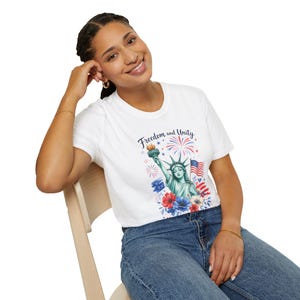 May include: A white short-sleeve t-shirt featuring a graphic print of the Statue of Liberty, an American flag, fireworks, and red, white, and blue flowers. The text Freedom and Unity is printed in dark blue script above the illustration.