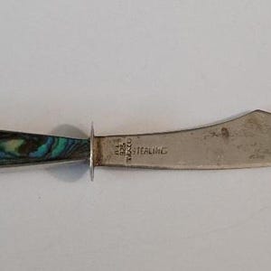 May include: A silver letter opener with a curved blade and a handle inlaid with iridescent abalone shell. The blade is stamped with the text 925 MEXICO STERLING. The handle features a circular abalone inlay at the end and a long tapered inlay.
